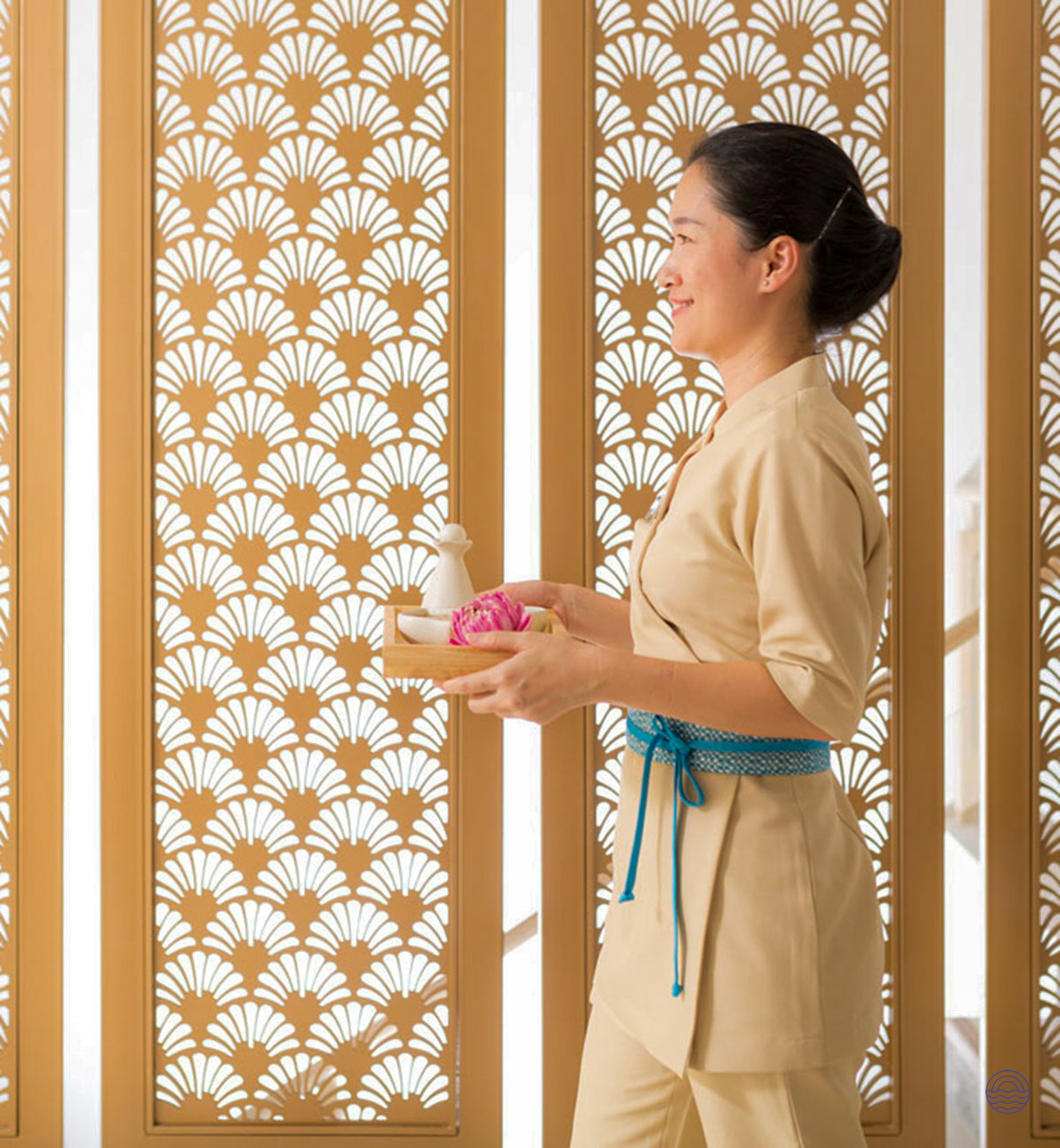 A smiling spa therapist in Phuket carries a tray with luxurious treatments in a serene setting.