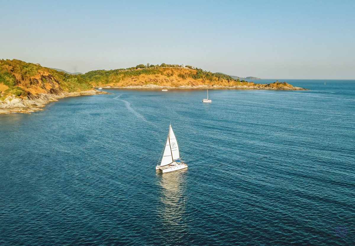 A white catamaran sails on the blue Andaman Sea near the green coast of Phuket.