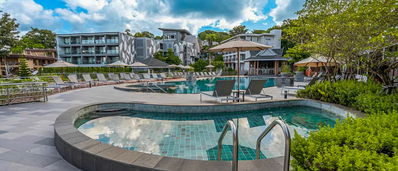 A spacious resort swimming pool area in Phuket with multiple pools, lounge chairs, and modern buildings.
