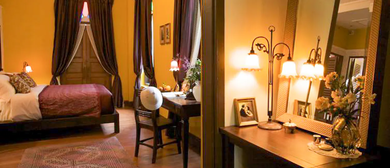 Warmly lit hotel room in Bangkok with a comfortable bed, wooden desk, and elegant mirror.