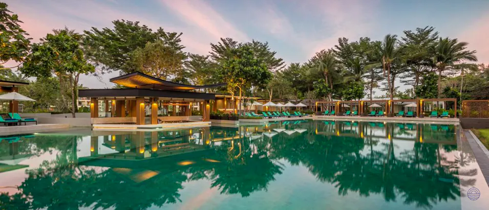 Luxurious resort swimming pool and bar area surrounded by tropical trees at sunset in Thailand.