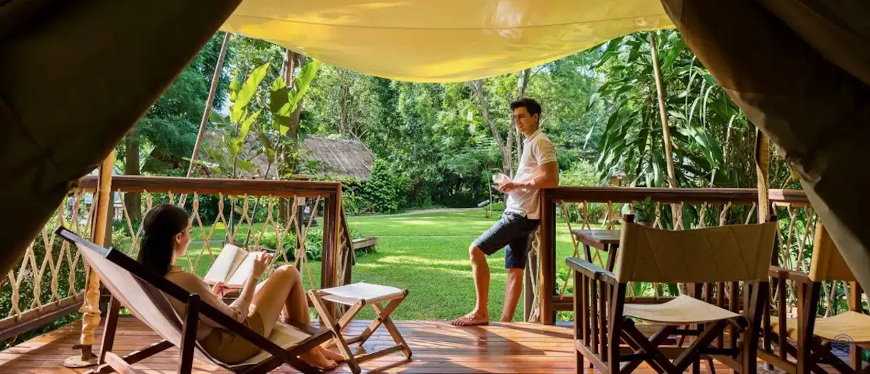 A couple relaxes on a wooden deck outside their glamping tent in lush Kanchanaburi, Thailand.