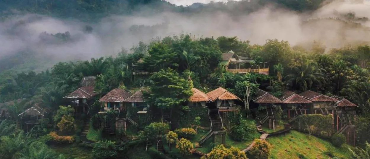 Tranquil bungalows nestled on a lush green hillside with misty mountains in Khao Sok, Thailand.