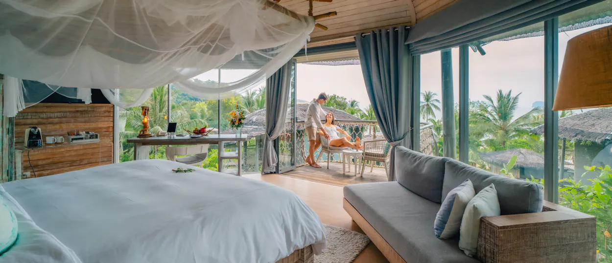 A couple relaxes on a balcony with tropical jungle views from their luxurious Koh Yao Noi room.