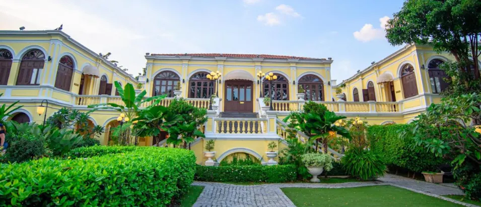 Elegant yellow colonial-style hotel with grand entrance and lush tropical gardens in Thailand.