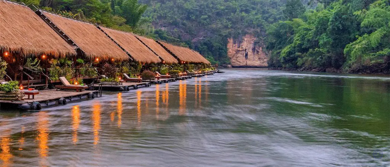 Charming thatched bungalows with warm lights float on the peaceful River Kwai in Kanchanaburi, Thailand.