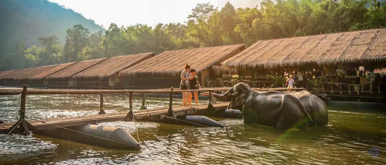 A couple interacts with elephants in a river next to floating bungalows in Kanchanaburi, Thailand.