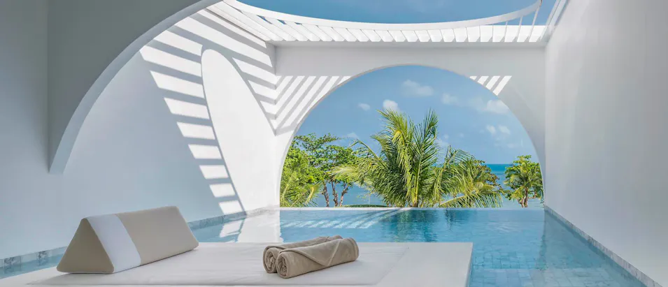 A modern white private pool villa overlooking the ocean with tropical palm trees and blue sky.