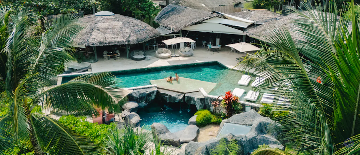 Two people relaxing in a stunning tropical resort pool with waterfall and palm trees on Koh Yao Noi.