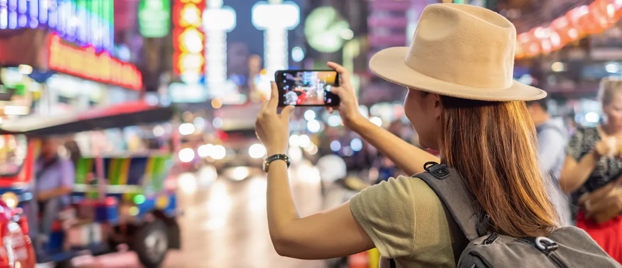 A traveler in a hat photographs the bustling, neon-lit streets and traffic of Bangkok at night.