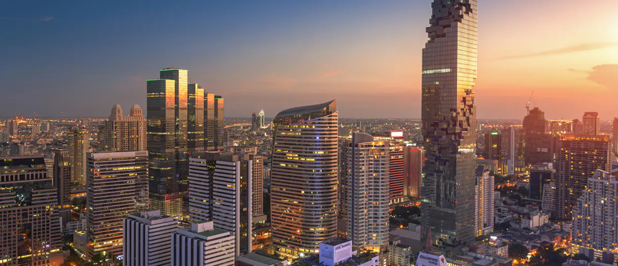 An aerial view of Bangkok's modern skyline at sunset, with illuminated skyscrapers and a vibrant sky.