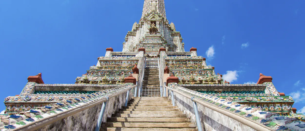 Ornate stairs ascend towards the majestic, towering Wat Arun temple in Bangkok under a blue sky.