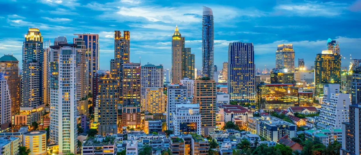 A vibrant Bangkok cityscape at dusk, with numerous illuminated skyscrapers against a deep blue sky.