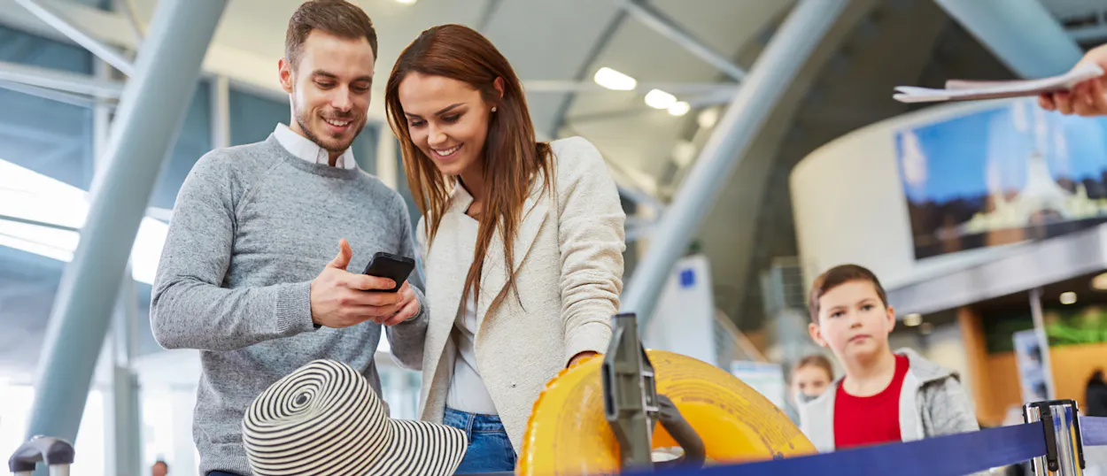 A smiling couple looks at a phone at the airport, with luggage ready for their trip.