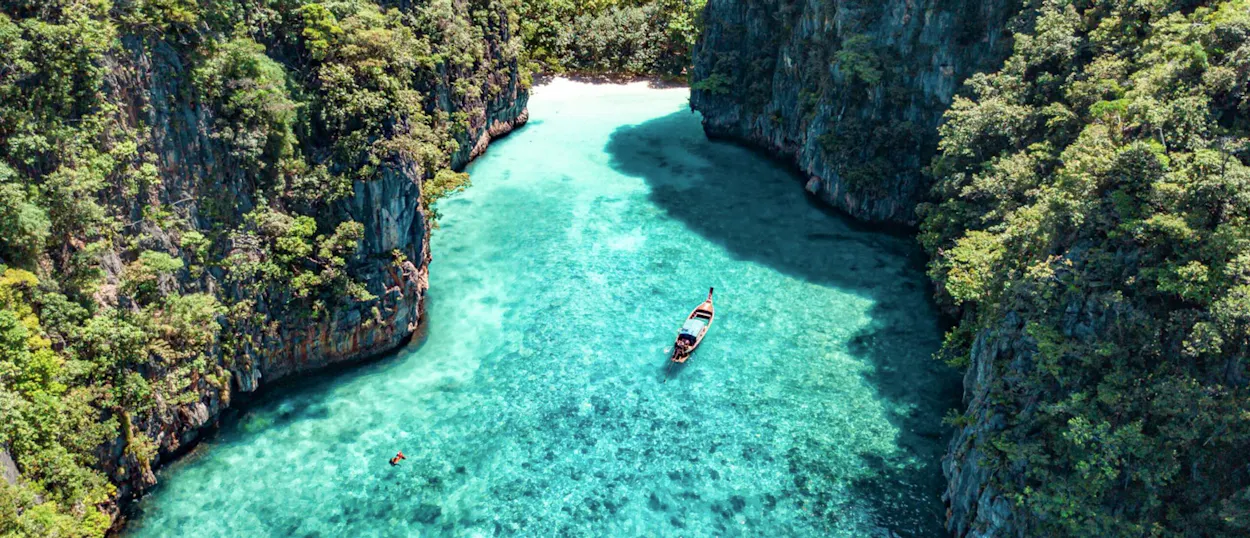 A longtail boat floats in crystal-clear turquoise waters surrounded by lush green cliffs in Koh Phi Phi.