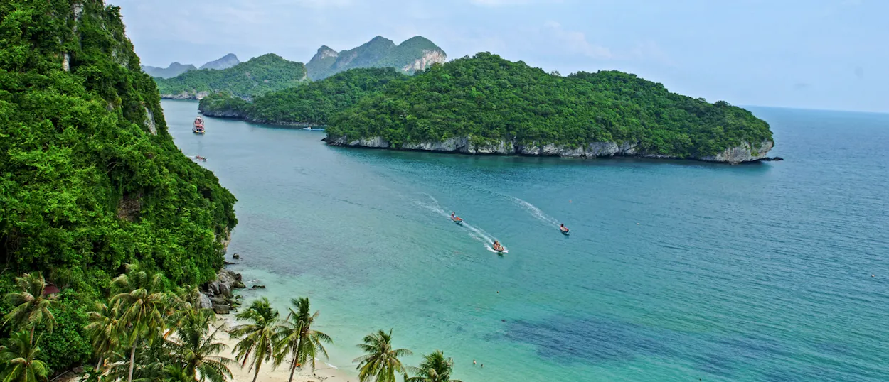 Boats cruise past lush green islands and a sandy beach with palm trees in Koh Samui.