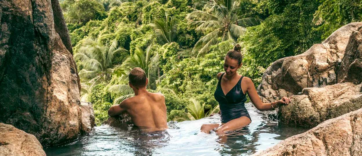 A couple relaxes in a natural rock pool overlooking a lush jungle in Koh Samui.