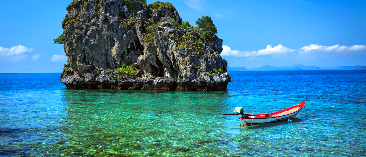 A red longtail boat floats on clear turquoise water beside a lush rocky island in Krabi, Thailand.