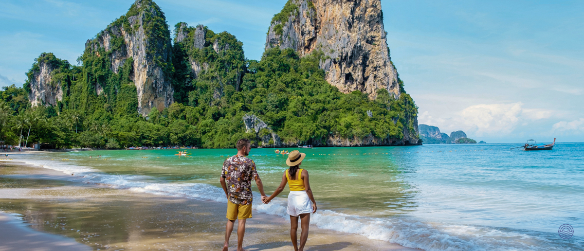 A couple walks hand-in-hand on a sandy Krabi beach, with towering green limestone cliffs and blue water.