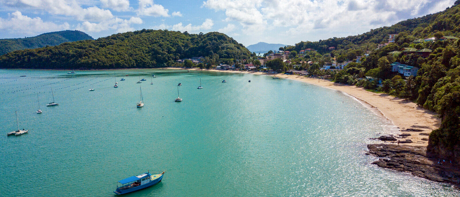 Aerial view of a serene tropical bay with turquoise water, sandy beach, and anchored boats in Phuket.