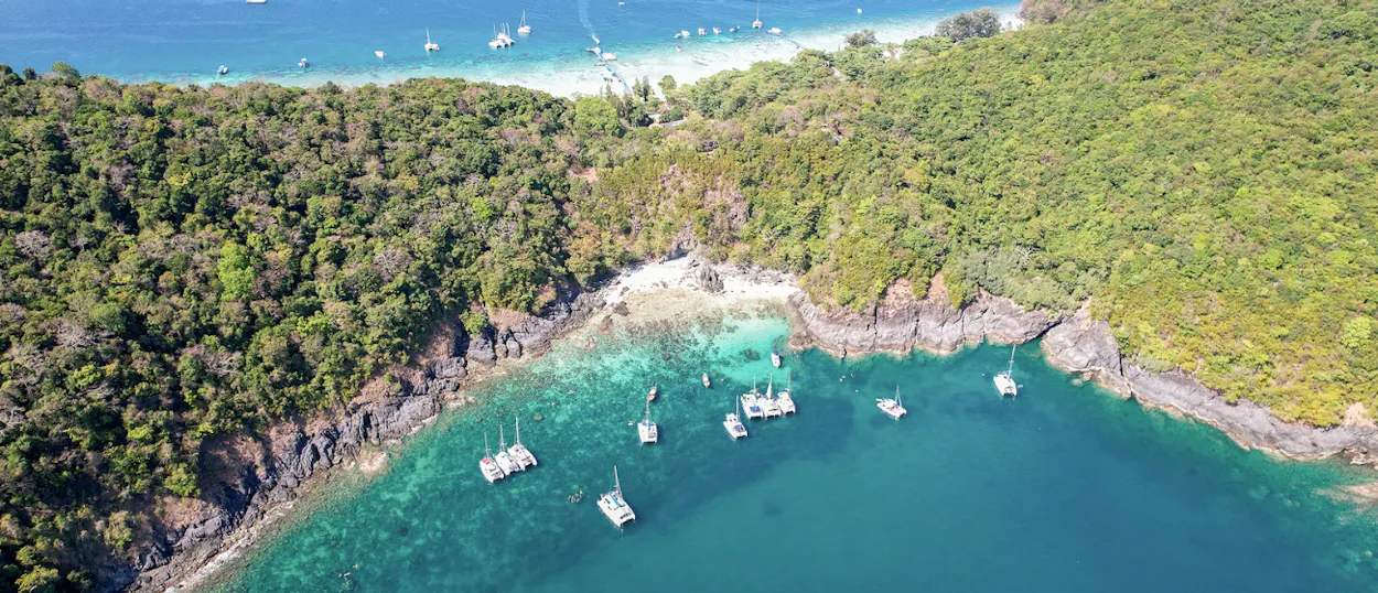 Lush green tropical island in Phuket with clear turquoise water and many anchored boats.