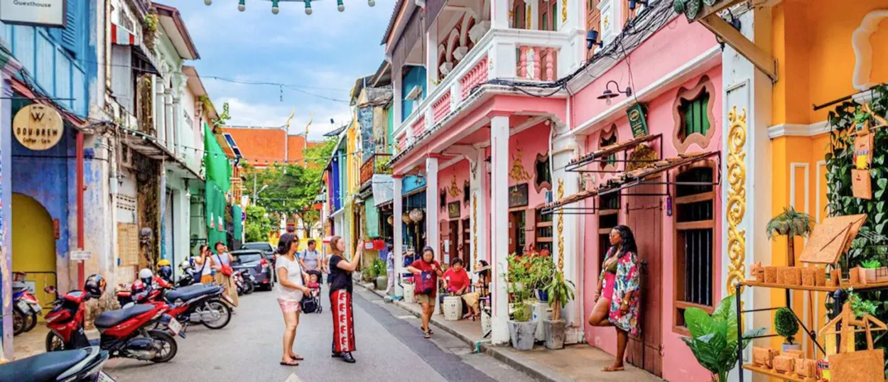 Tourists explore a vibrant street lined with colorful Sino-Portuguese buildings in Phuket Old Town, Thailand.