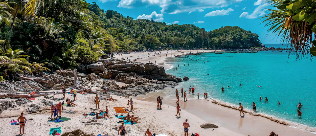 Many people swim and relax on a beautiful sandy Phuket beach with turquoise water and green hills.