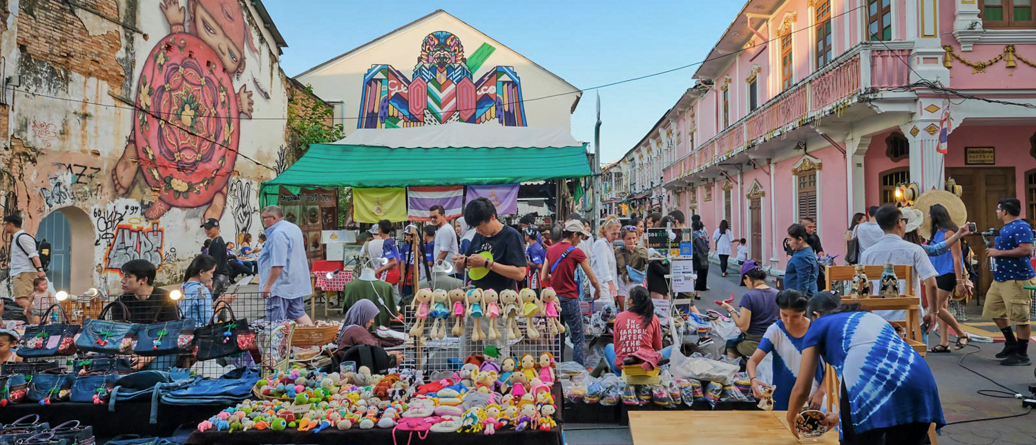 A bustling street market in Phuket Old Town with people browsing colorful stalls and unique street art.
