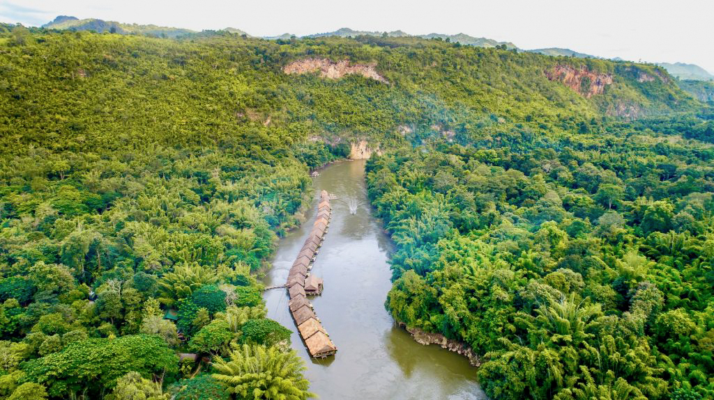 Floating bungalows line the tranquil River Kwai, surrounded by dense green jungle and distant mountains.