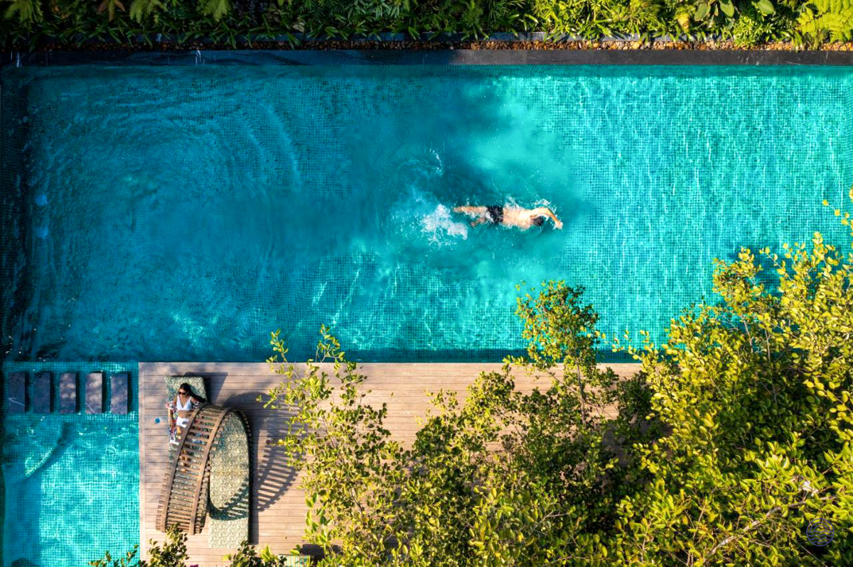 A person swims in a serene blue pool, another relaxes on a deck, surrounded by lush greenery.
