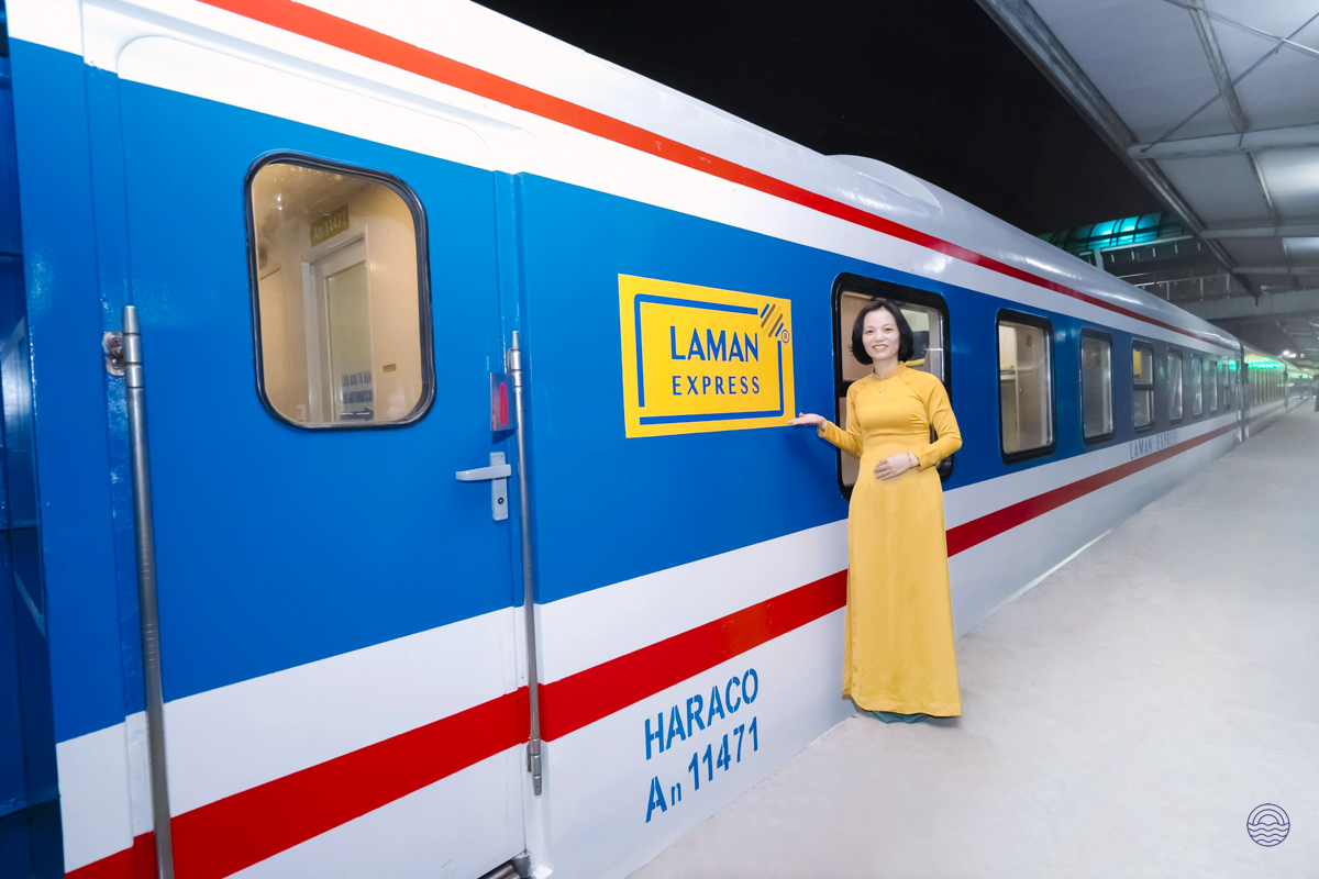 An elegant woman in a yellow ao dai stands beside the blue Laman Express overnight train.