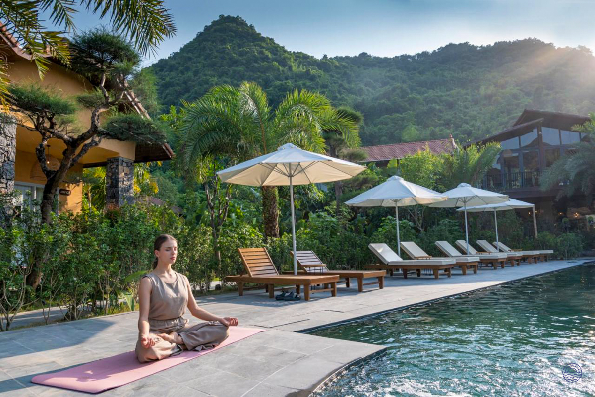 A woman meditates by the serene pool at Tam Coc Retreat, surrounded by lush tropical mountains.