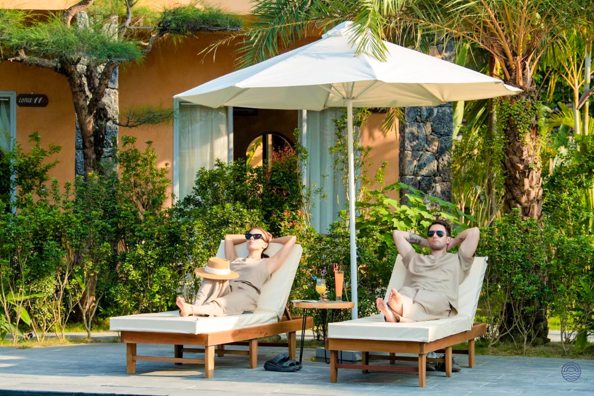 Two guests unwind on sun loungers under an umbrella amidst lush gardens at a Tam Coc retreat.