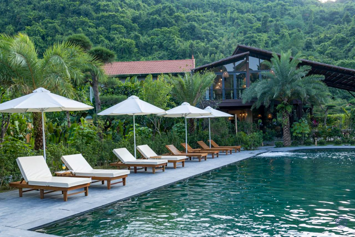 A tranquil swimming pool at Tam Coc Retreat, surrounded by lush tropical gardens and a forested mountain.