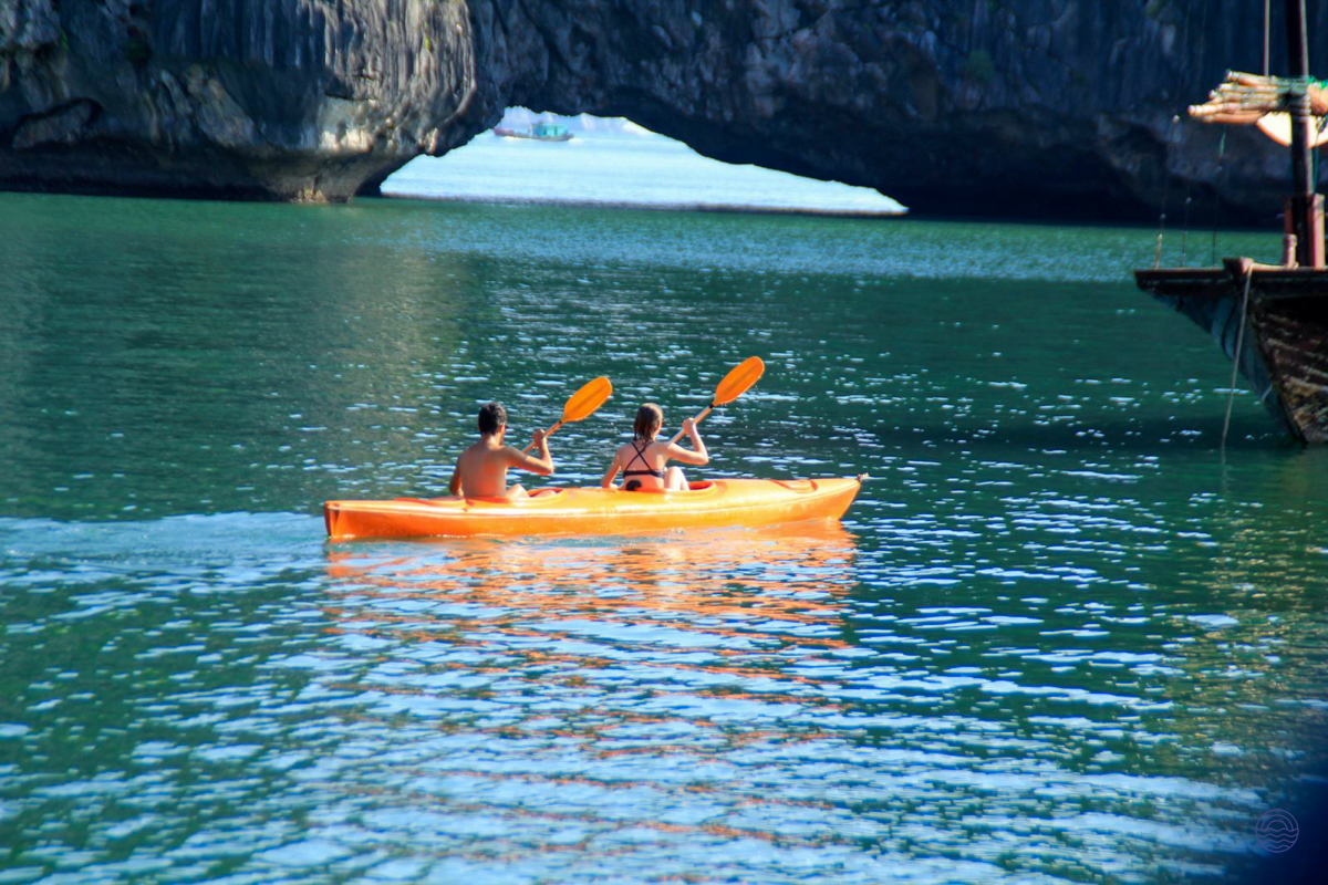 Two people paddle an orange kayak through the emerald waters of Halong Bay, past a natural arch.