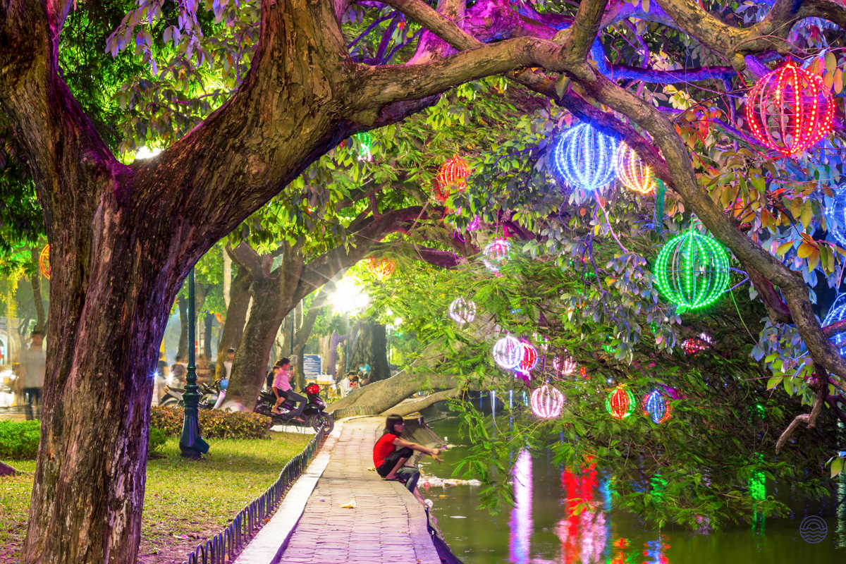 A person relaxes by the water under trees adorned with vibrant, colorful lanterns in Hanoi at night.