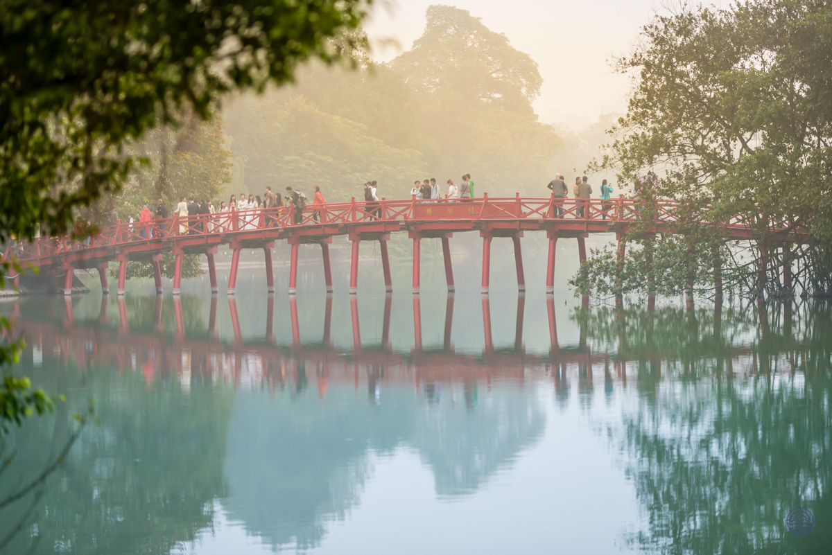 People stroll across the iconic red Huc Bridge, reflected beautifully in Hanoi's misty Hoan Kiem Lake.