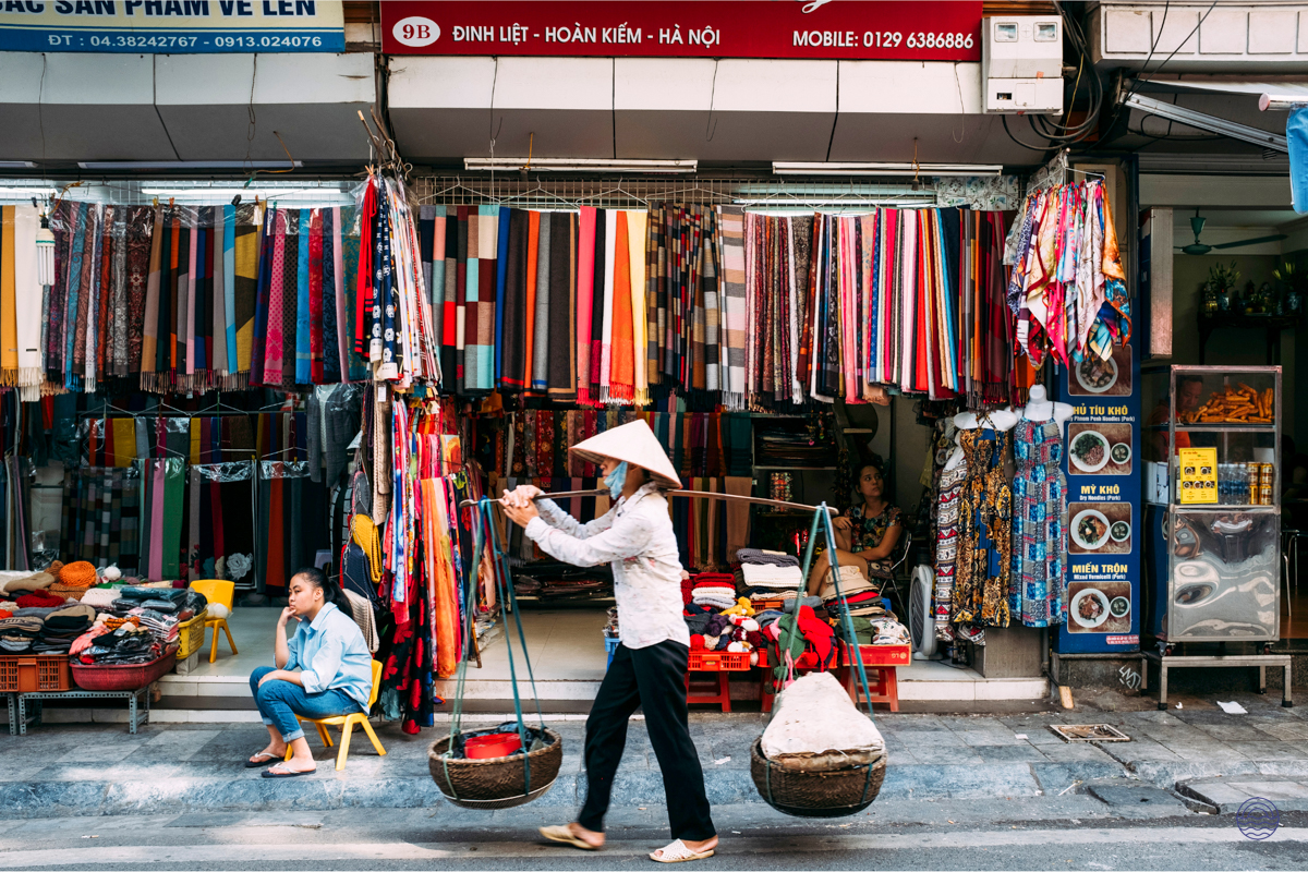 A street vendor in a traditional conical hat walks past colorful textiles in a bustling Hanoi market.
