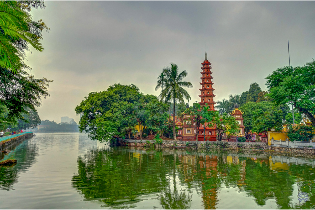 The historic red Tran Quoc Pagoda stands on West Lake in Hanoi, surrounded by lush green trees.