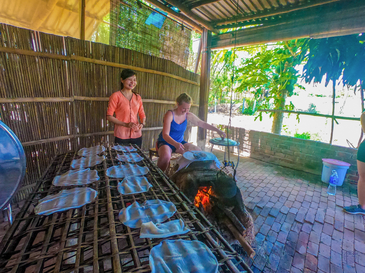 Two women learn traditional cooking techniques over an open fire in a rustic Hoi An kitchen.