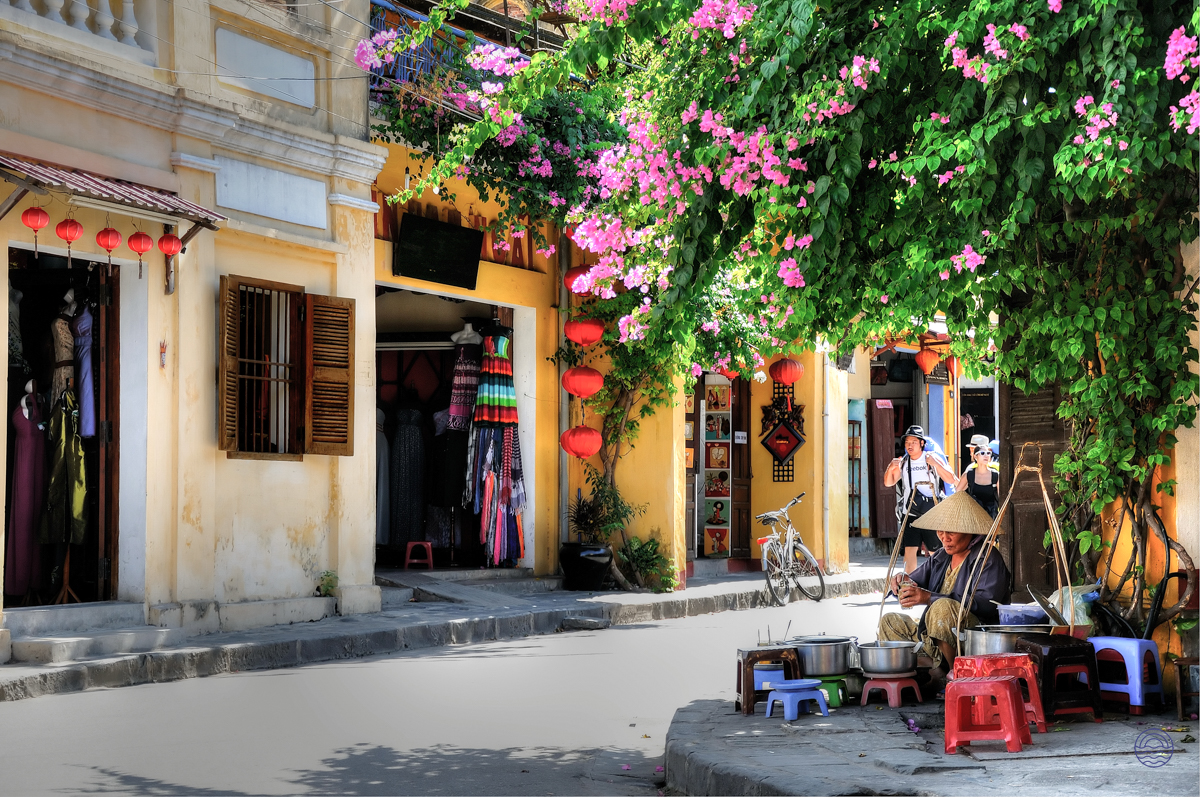 A street vendor prepares food on a charming street in Hoi An, beneath vibrant pink bougainvillea.
