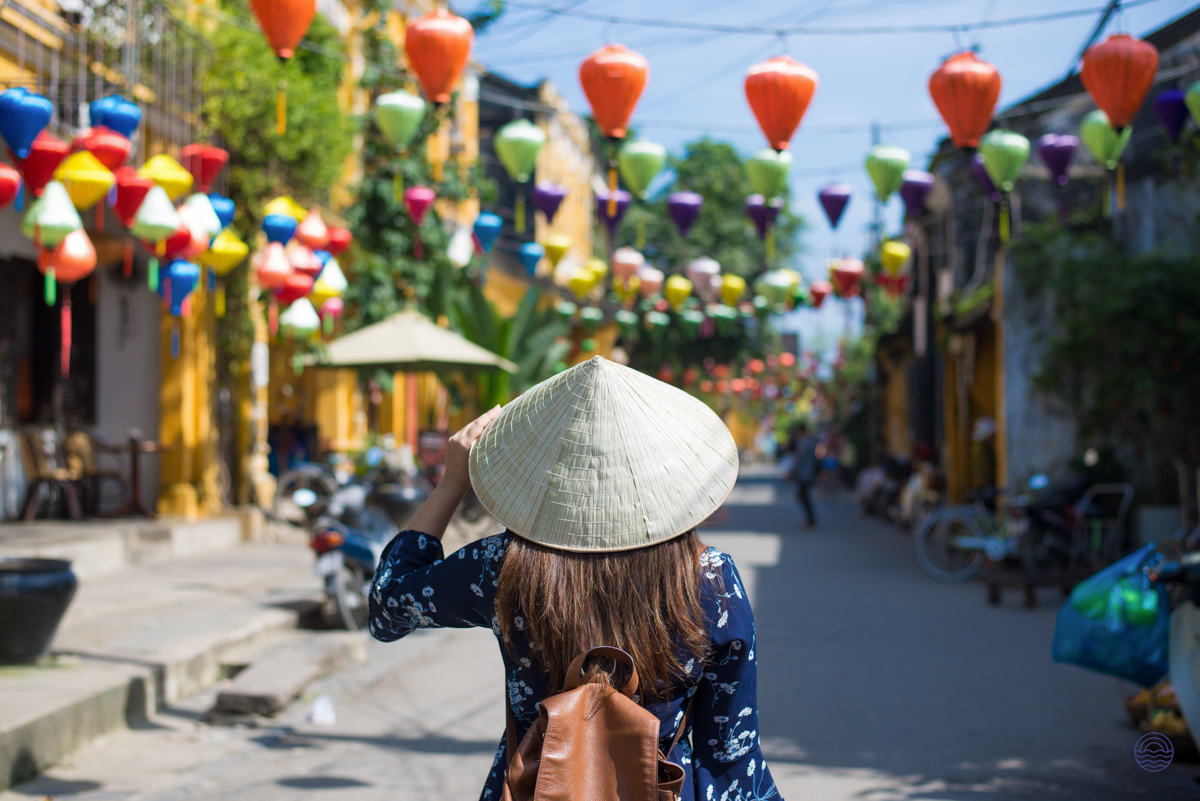 A person wearing a conical hat explores a vibrant street in Hoi An, adorned with colorful lanterns.