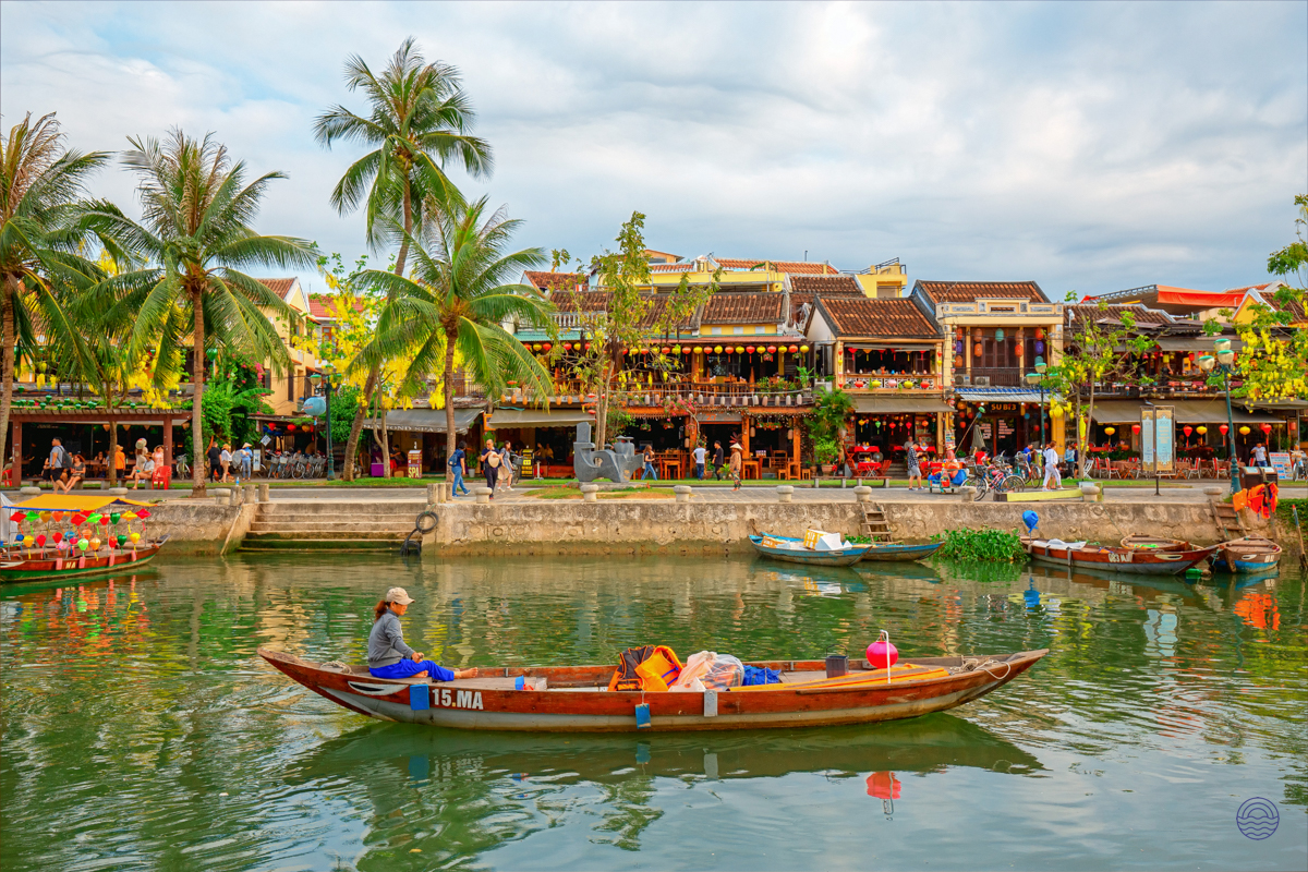A traditional boat glides on the serene river in Hoi An, past charming buildings and swaying palm trees.
