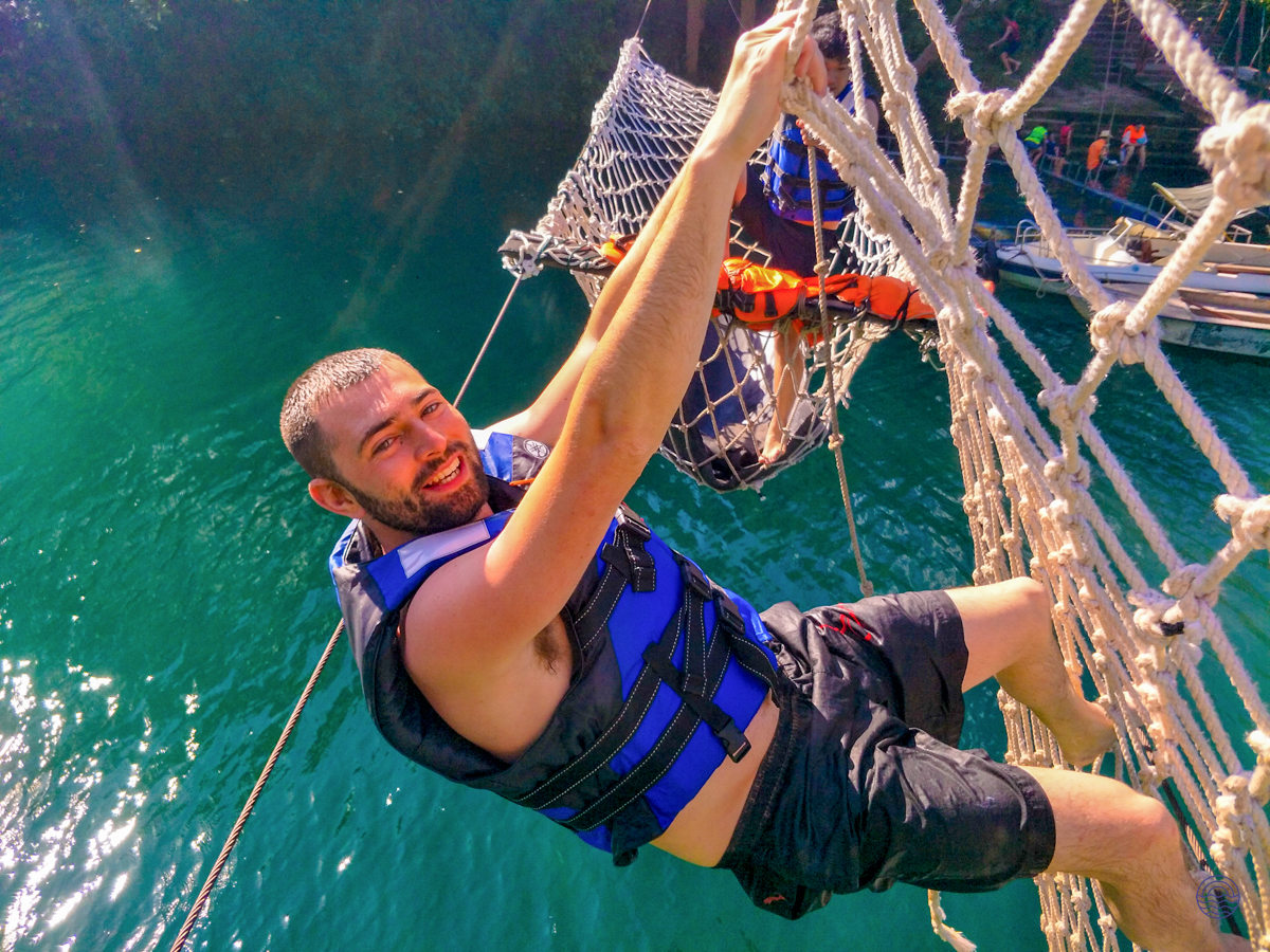 A smiling man in a life vest climbs a rope net over the turquoise waters of Phong Nha.