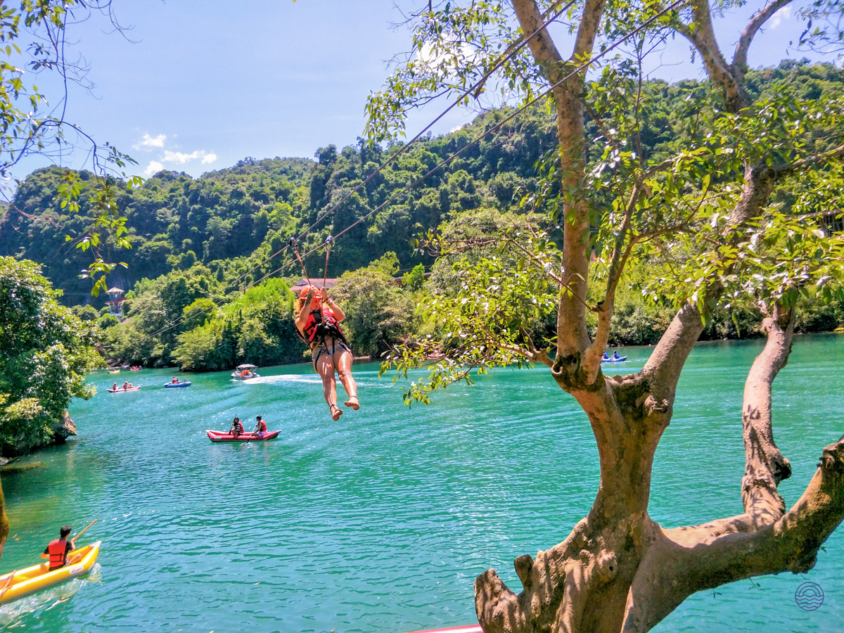 A person zip-lines over the clear turquoise river in Phong Nha, surrounded by lush green mountains.
