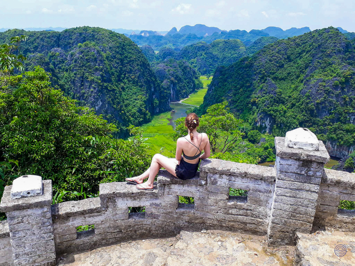 A woman sits on a stone wall, admiring Tam Coc's breathtaking landscape of green karsts and winding river.