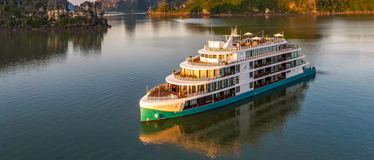 Elegant Capella cruise ship glides past limestone islands in Halong Bay during golden hour.