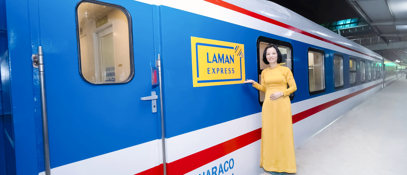 Smiling woman in yellow ao dai stands beside the blue Laman Express train in Vietnam.