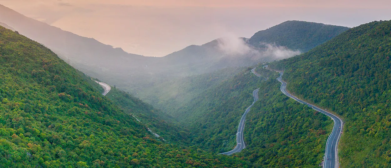 Winding roads snake through lush green mountains with misty valleys at Hai Van Pass.
