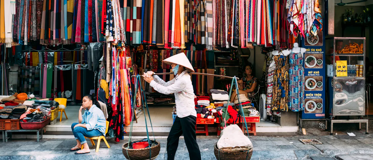 A street vendor in a conical hat carries baskets past colorful fabric stalls in Hanoi.